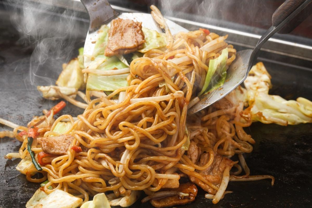 Yakisoba noodles with meat and vegetables being cooked on griddle with metal spatula