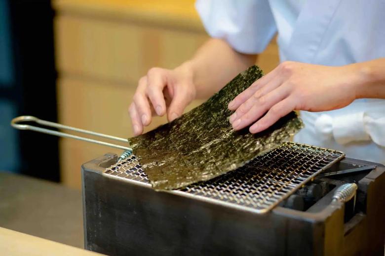 Hands toasting nori seaweed sheet over flame with metal grater