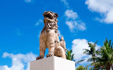 Stone lion statue on white pedestal against blue sky with palm trees