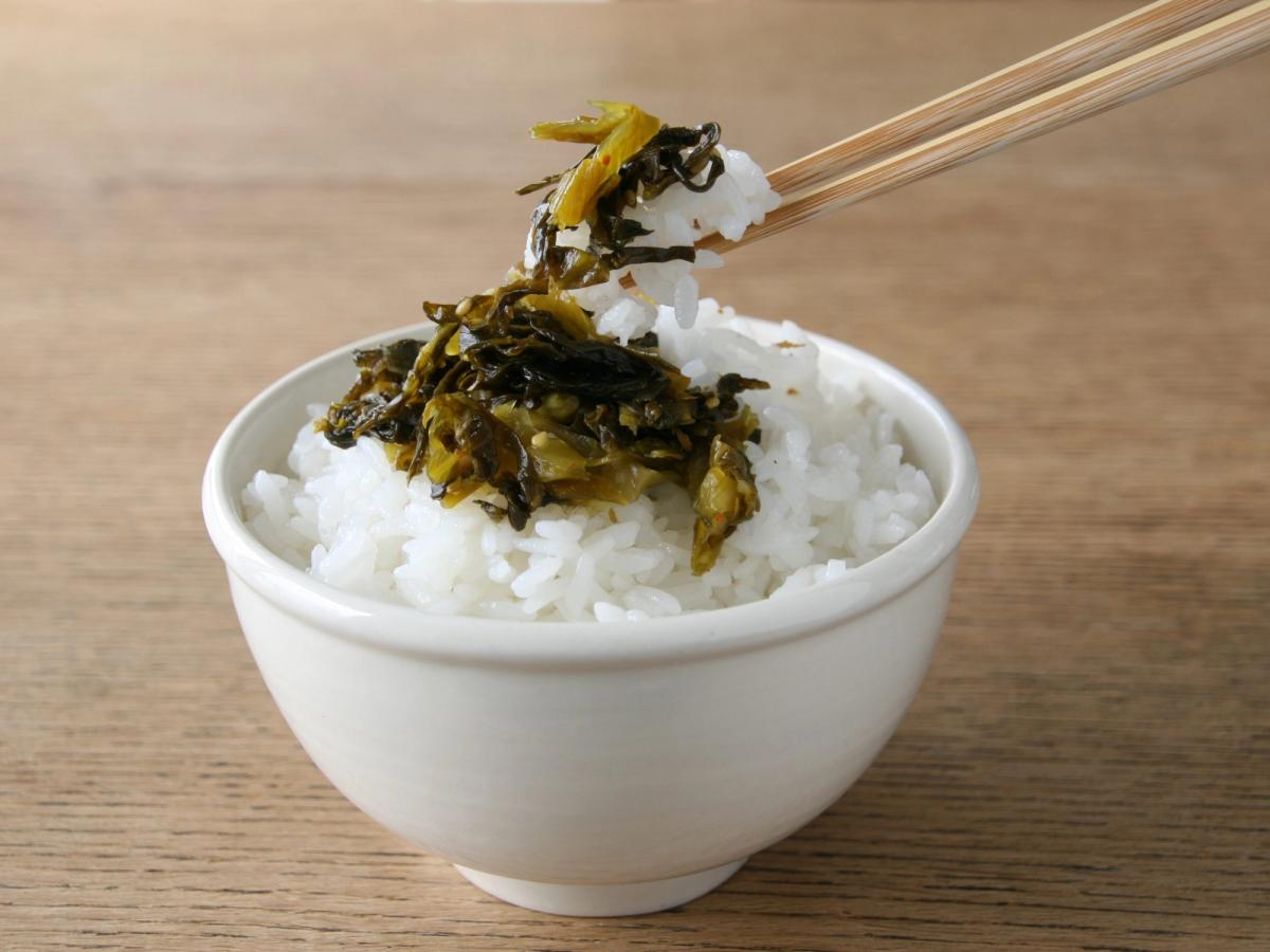 Chopsticks lifting pickled mustard greens over white rice in ceramic bowl