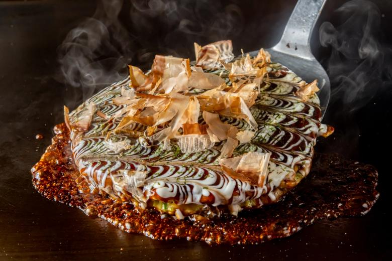 Okonomiyaki with decorative drizzle pattern and bonito flakes steaming on dark griddle