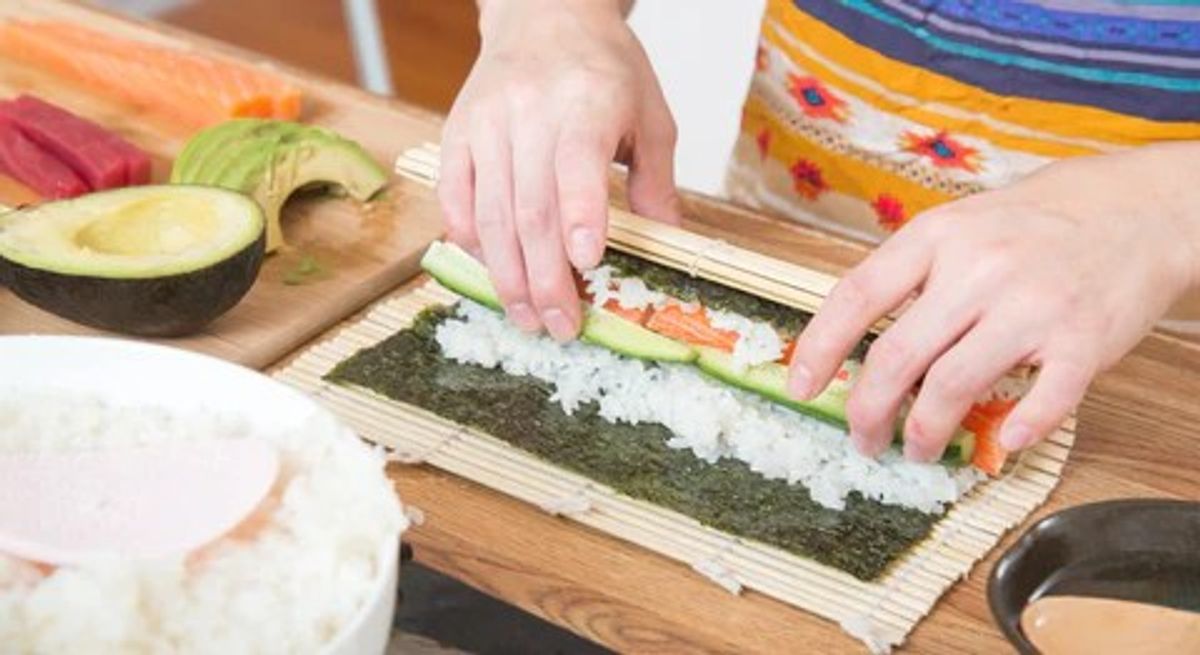 Person rolling sushi with nori, rice, avocado, and vegetables on bamboo mat