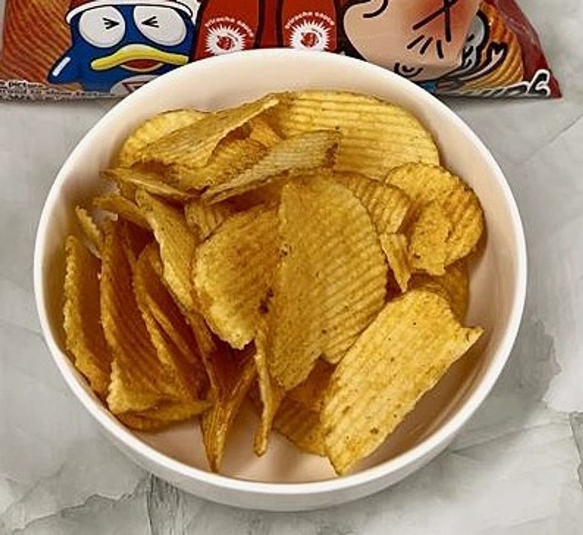 White bowl of golden ridged potato chips with snack package in background