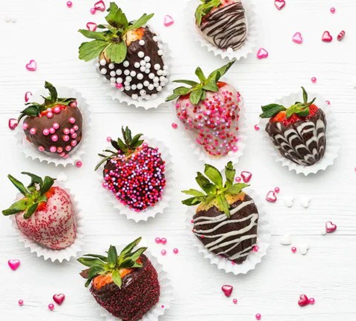 Chocolate-covered strawberries with decorative toppings and pink heart confetti on white background