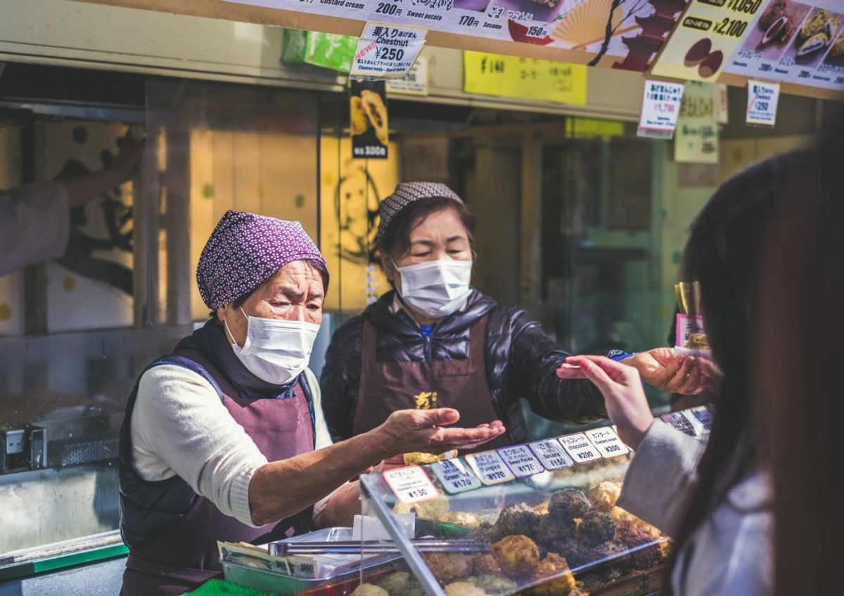 Two masked vendors serve customer at Japanese food stall counter with displayed dishes