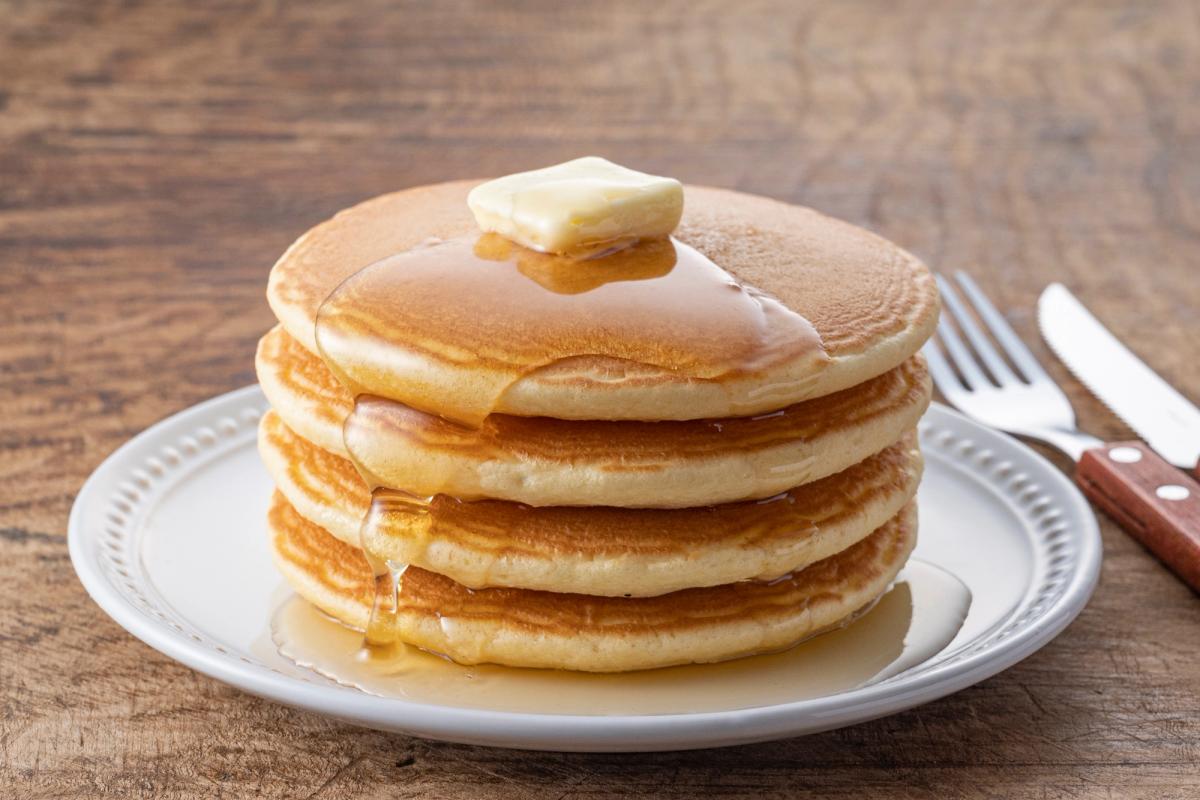 Stack of four fluffy pancakes with butter and syrup on white plate, wooden table background