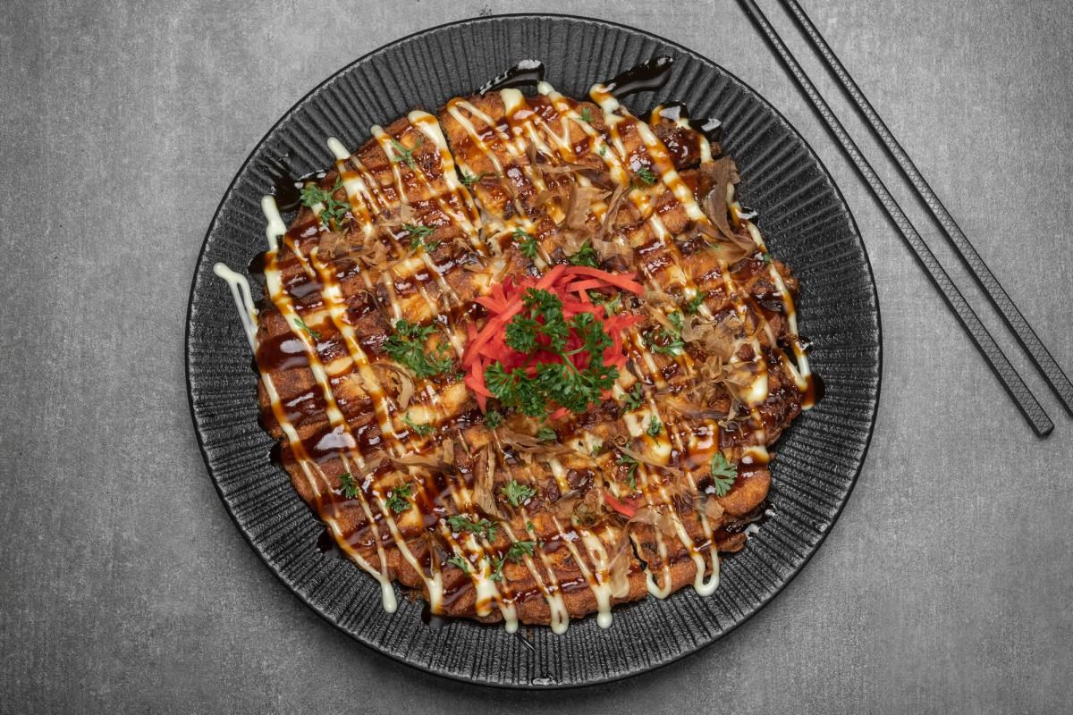 Okonomiyaki topped with sauce, mayo drizzle, pickled ginger, and green onions on black plate