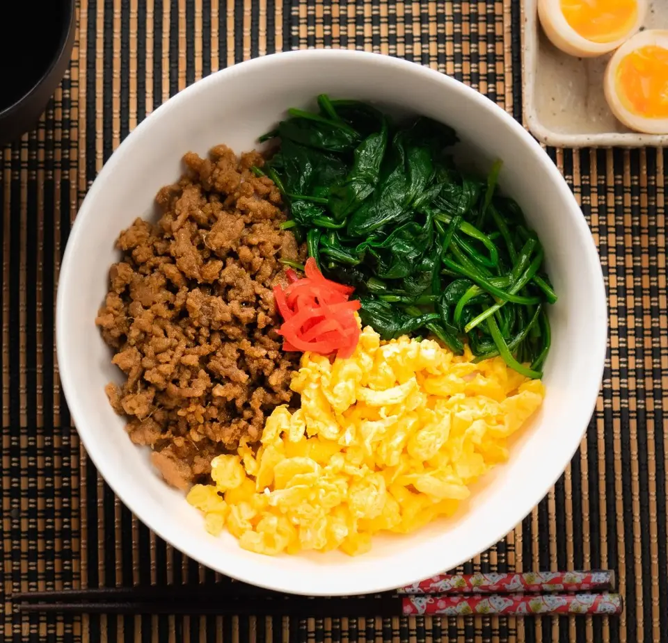 Bibimbap bowl with ground meat, spinach, egg, pickled radish, and rice on woven placemat