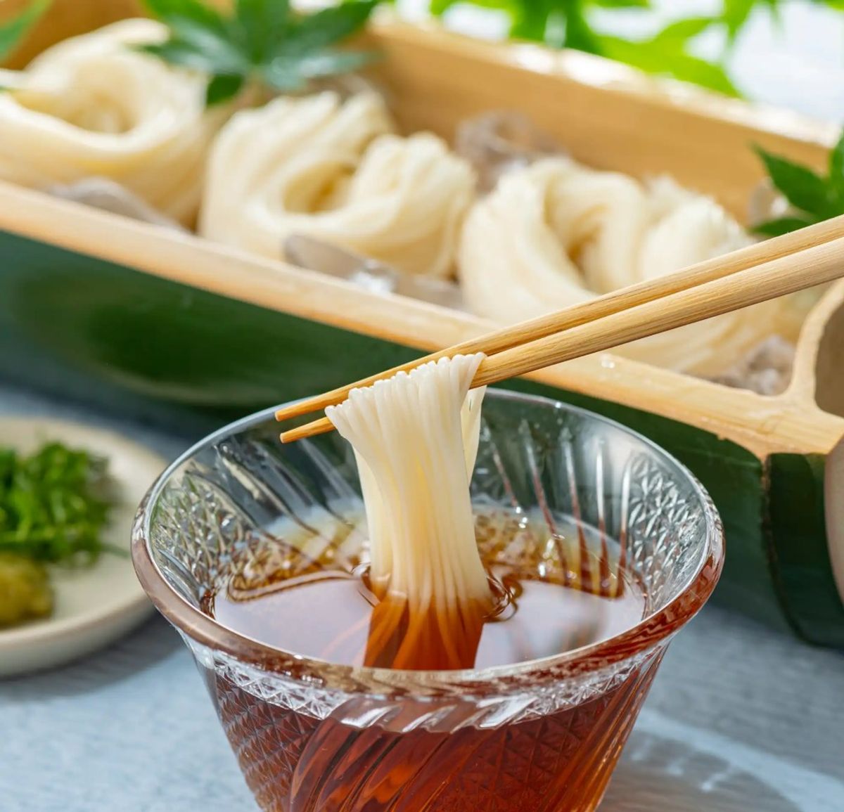Chopsticks holding noodle strand over dipping sauce bowl with dumplings in bamboo steamer background
