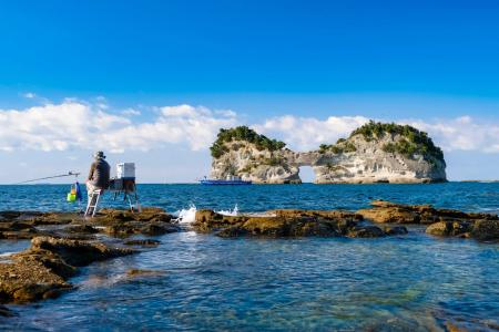 Fisherman sitting on rocks casting line toward island with blue ocean and clear sky