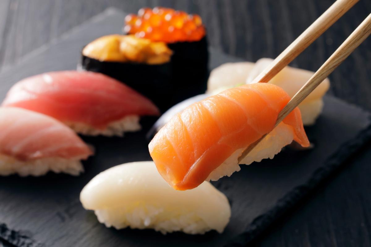 Assorted nigiri sushi on black slate with chopsticks holding salmon piece
