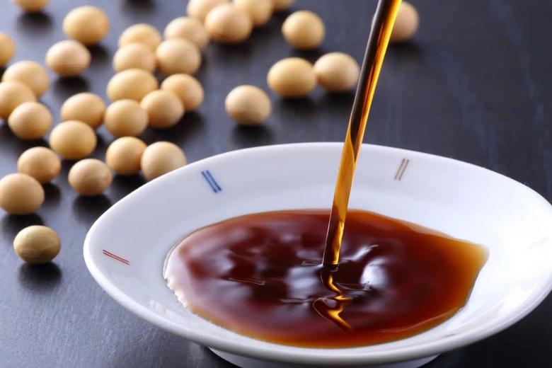 Soy sauce pouring into white bowl with soybeans scattered on dark surface