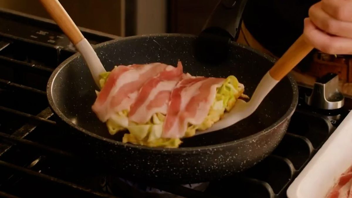 Bacon strips being placed on omelette in nonstick pan on stovetop