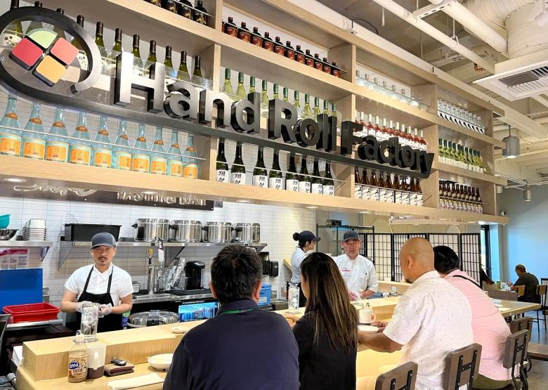Hand Roll Factory interior with sushi counter, staff, customers, and shelved beverages and ingredients