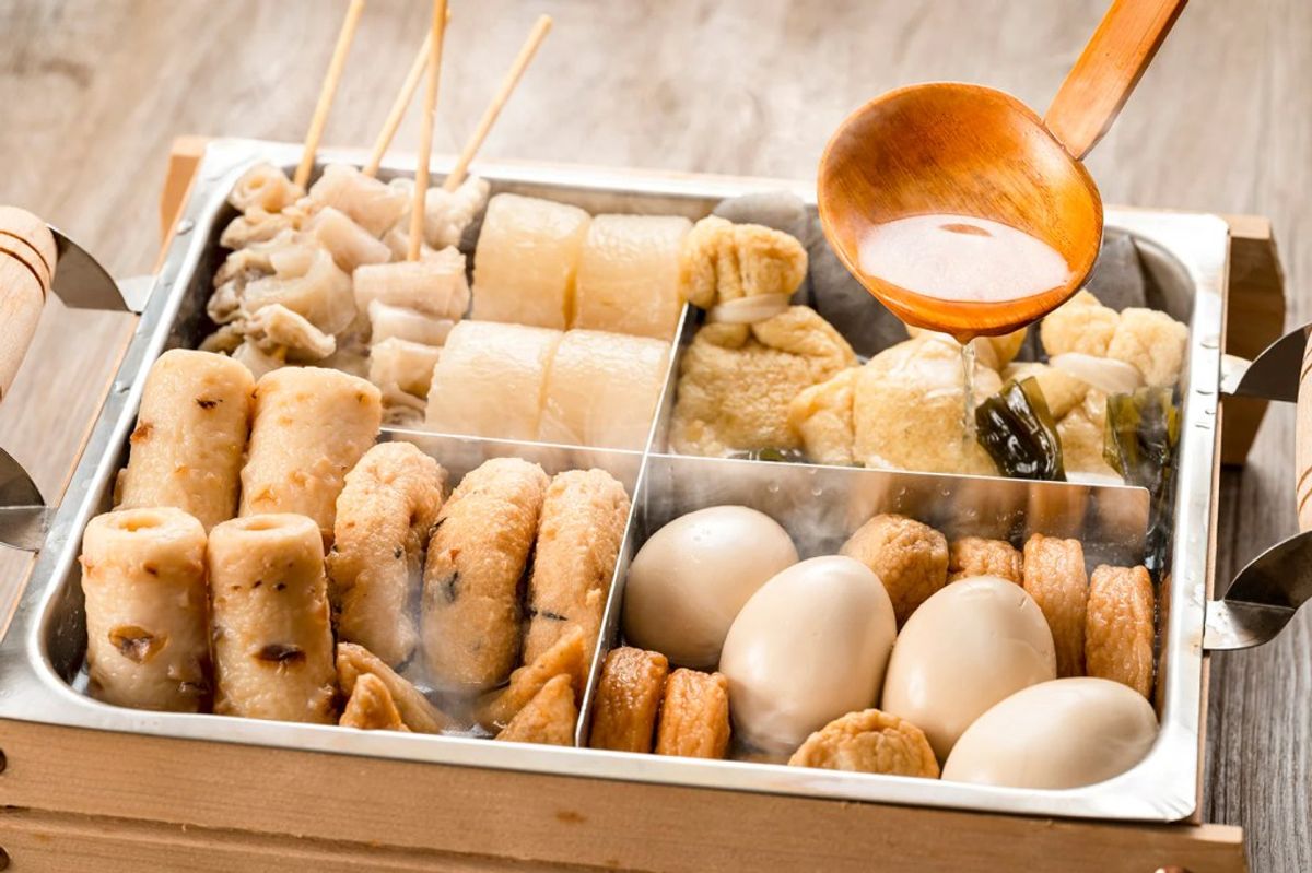 Assorted oden ingredients in compartmentalized tray with dipping sauce and skewers