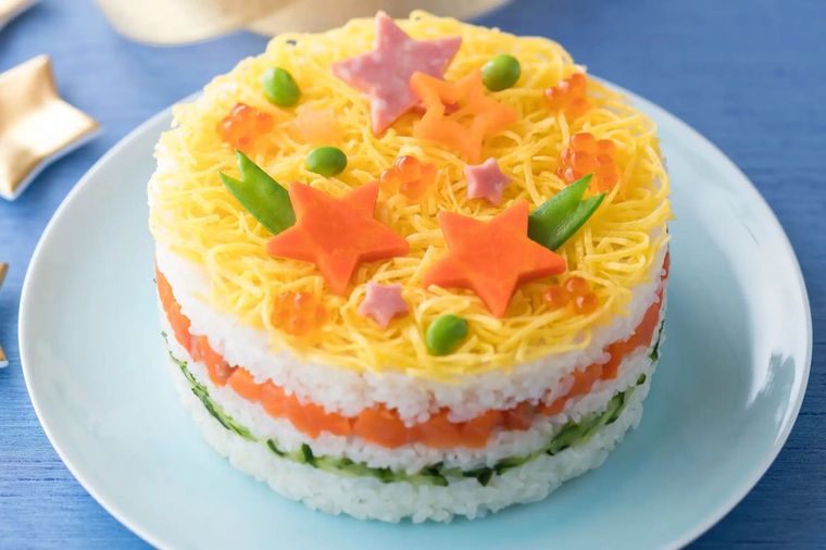 Colorful layered sushi cake with star-shaped toppings and vegetable decorations on white plate